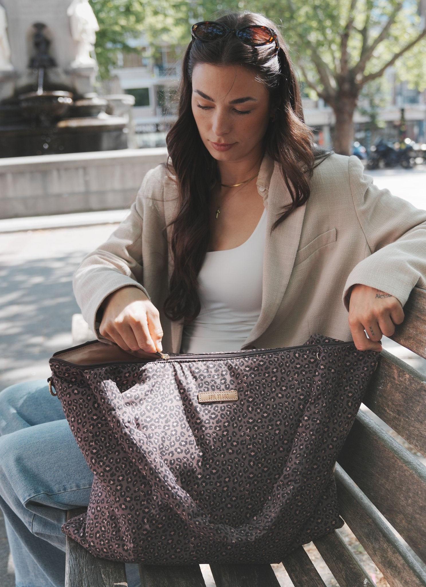 The Lily Tote luiertas van Love by Lily in Wild Espresso luipaardprint, op een bankje terwijl iets in de tas wordt gestopt bij een fontein op een plein