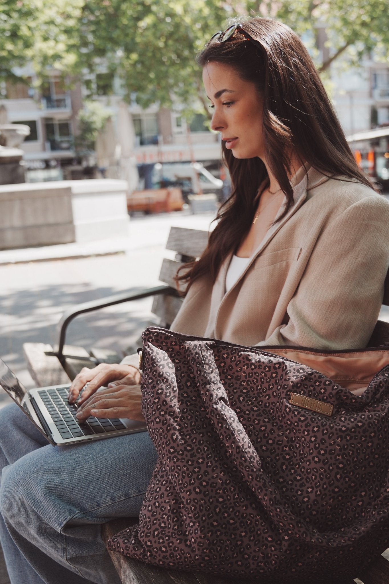 The Lily Tote luiertas van Love by Lily in Wild Espresso luipaardprint, naast een laptop op schoot op een bankje in de stad