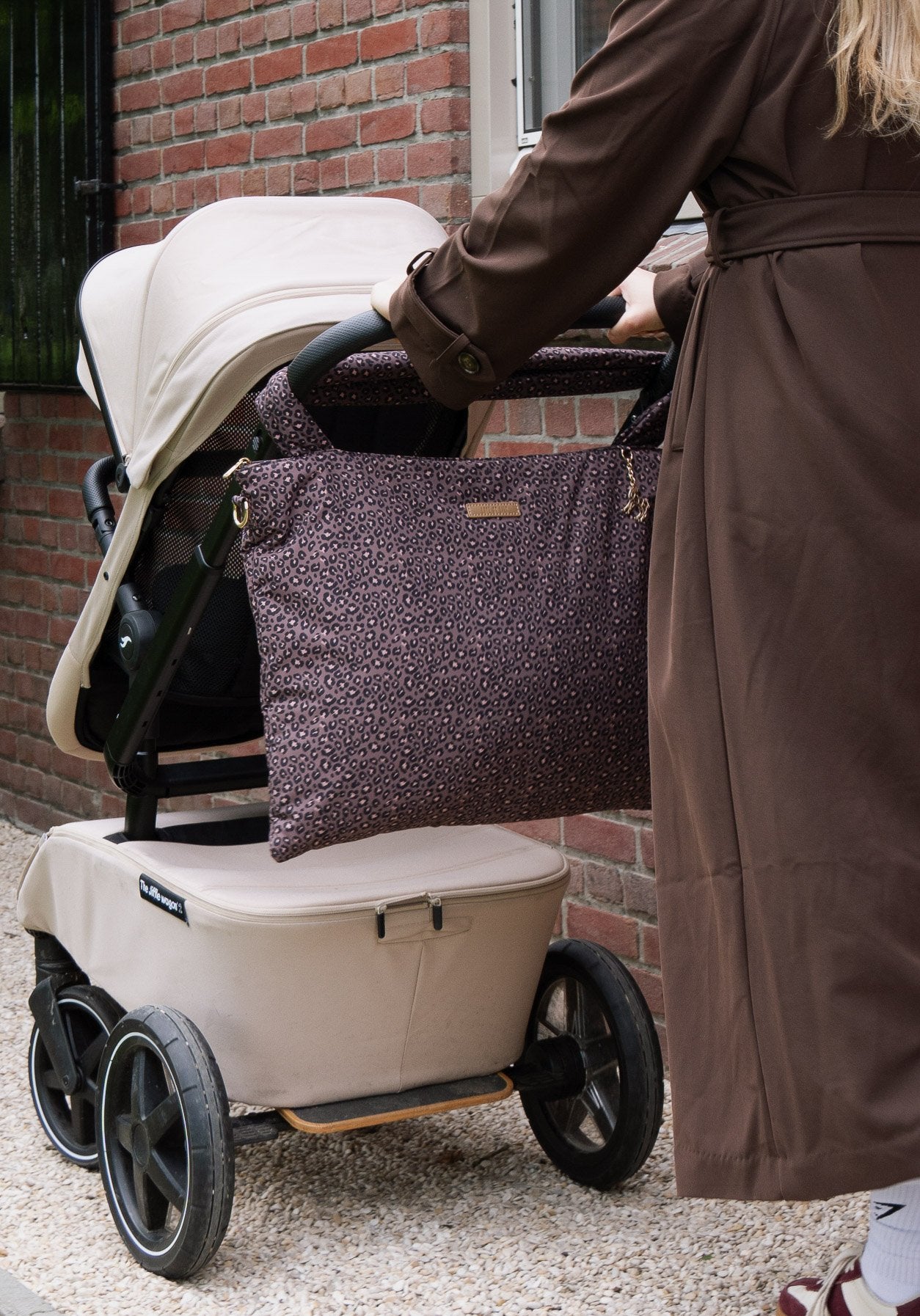 The Lily Tote luiertas van Love by Lily in Wild Espresso luipaardprint, hangend aan het duwhandvat van de kinderwagen terwijl een moeder in trenchcoat wandelt langs een bakstenen huis